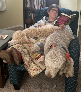 Kara sits on an armchair, two poodles in her lap and a book open in her hand.