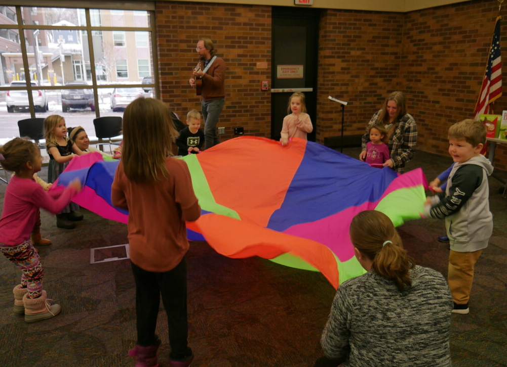 Pete playing guitar and singing while kids hold the sides of a multicolor parachute lifting it up and down to the song.