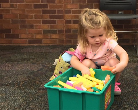 Small girl with a bag on her shoulder digs through bin of toys.
