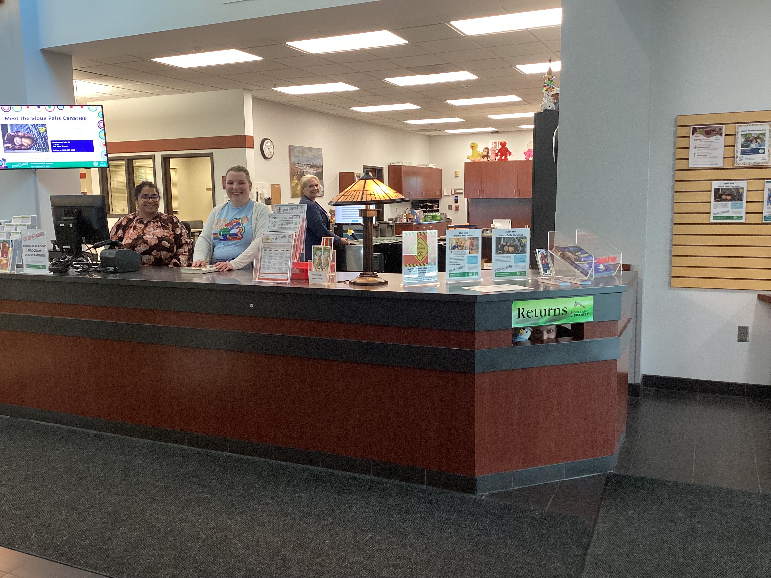 Staff at the front desk smile at the camera, while a pair of eyes stares out from the Returns slot.