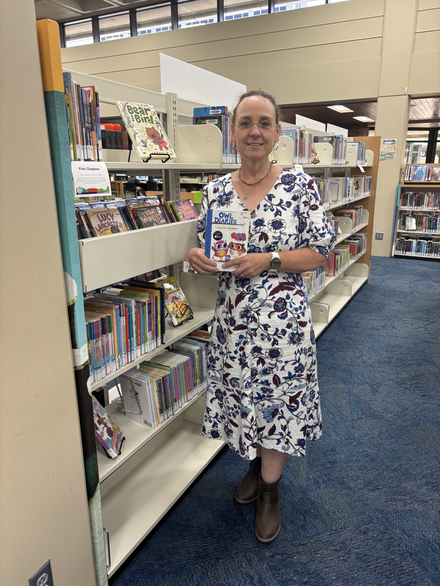 Library Associate wearing a white dress with blue floral pattern standing next to a shelf of the new First Chapters collection, holding one of the titles.