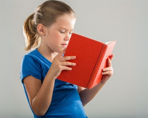 Female elementary student with dark blonde ponytail wearing a blue tshirt, holding and reading an open book with a red cover.