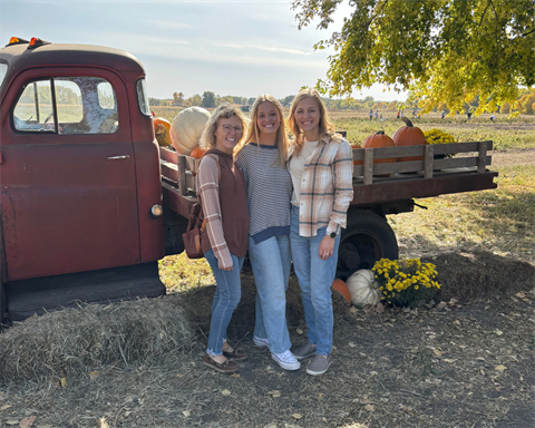 Author Leah T, her mother, and sister standing and smiling in front of an old red flatbed pickup full of pumpkins on a sunny fall day.