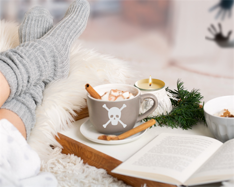 Person wearing grey comfy socks lounging on a plush pillow and blanket next to a serving tray with cocoa served in a grey mug with a skull and cross bones logo next to an open book