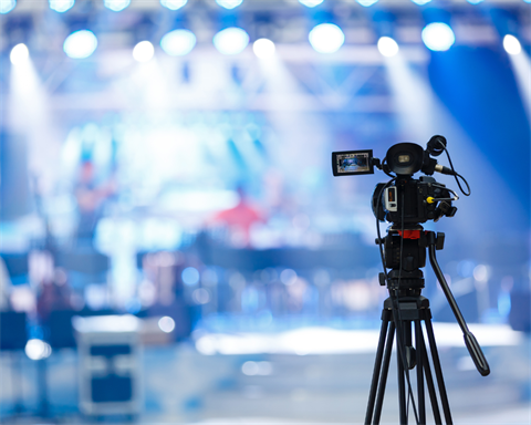 Video Camera mounted on a tripod in focus in the foreground pointed at an out of focus television production set with bright lights on stage rigging illuminating everything.