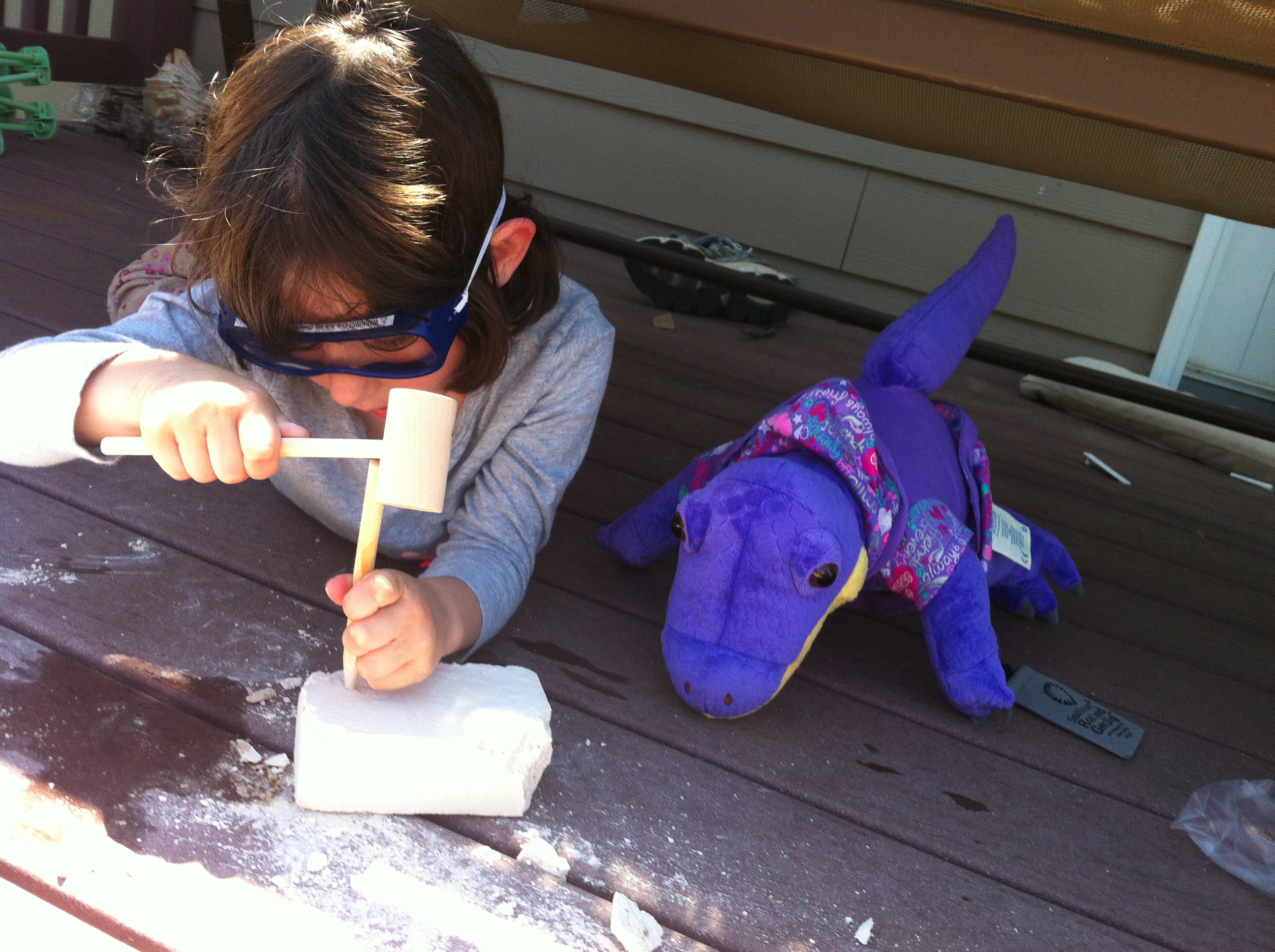 A young girl carves into stone, with a stuffed dinosaur beside her.