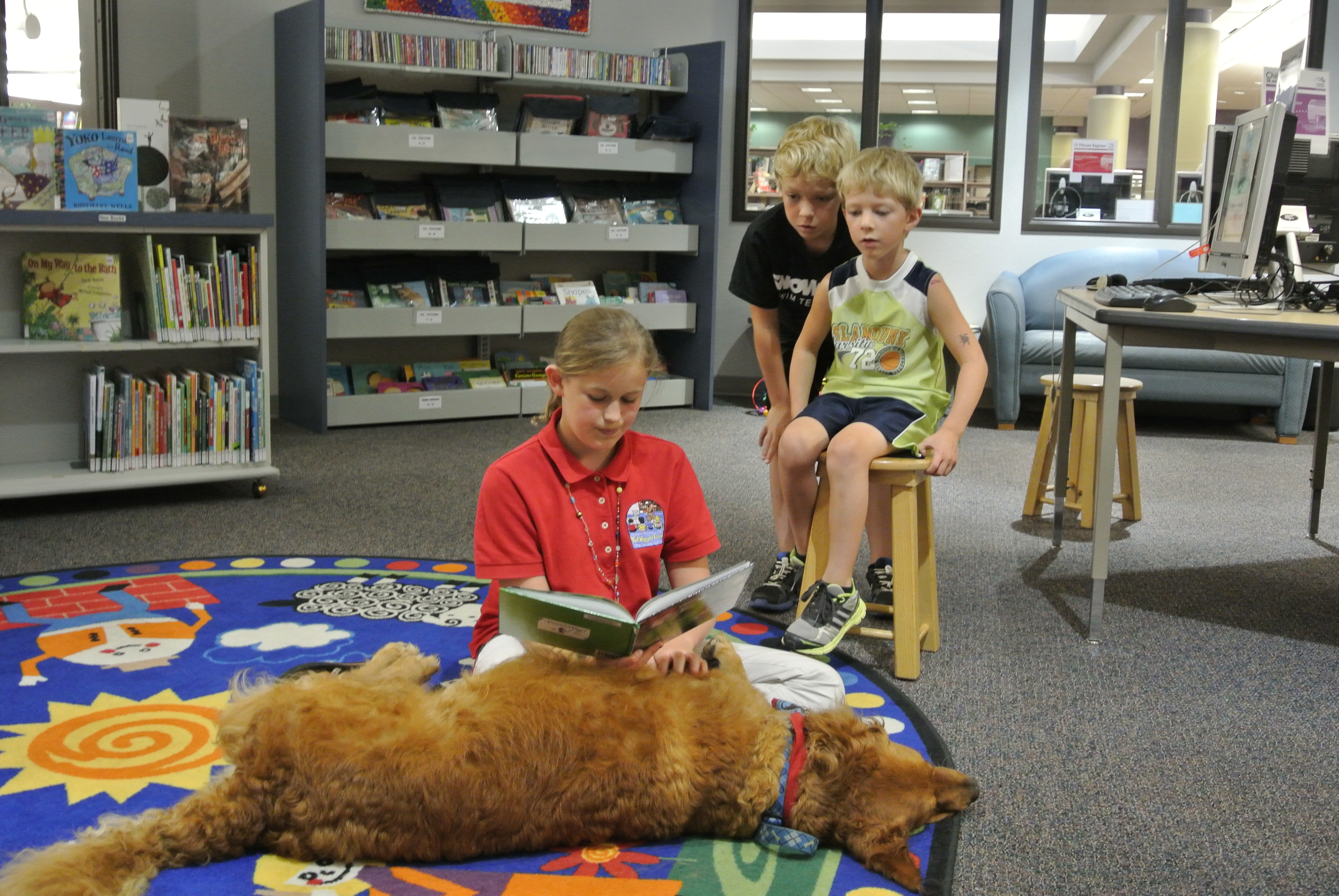 One child is seated on the floor with an open book next to a dog laying down on blue carpet with nursery rhyme characters printed on it. To children look on close by at the child reading to the tail waggin' tutor dog.