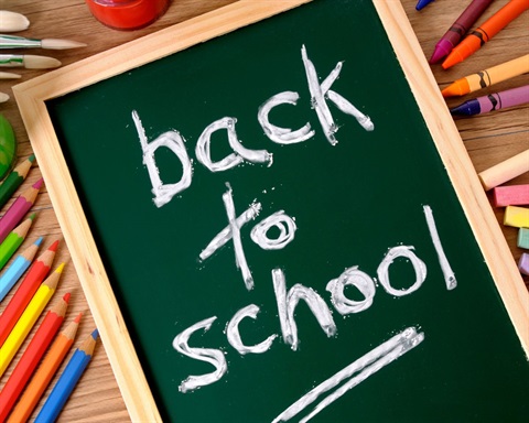 An angled, top-down photograph shows a green, wood-framed chalkboard centered on a light wooden table surface, surrounded by art and school supplies. Written in handwritten white chalk across three lines in the middle of the board is the text: back to school Underneath 