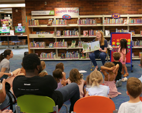 Children and families in the foreground are gathered facing a Siouxland Storyteller who is holding and reading a picture book for storytime in front of a wall of book shelves. A young child is standing next to the storyteller pointing to part of the storybook.