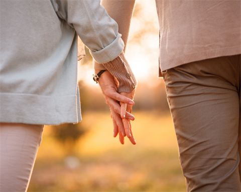 Close-up photograph of two people holding hands while walking outdoors together at sunset, bathed in warm, soft golden sunlight in the background.
