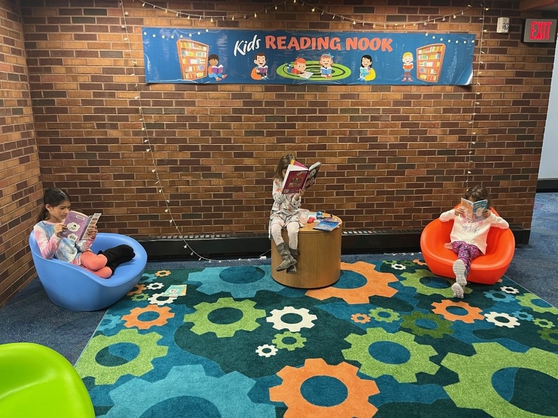 Three young girls are sitting in the library's children's area, each reading a book.