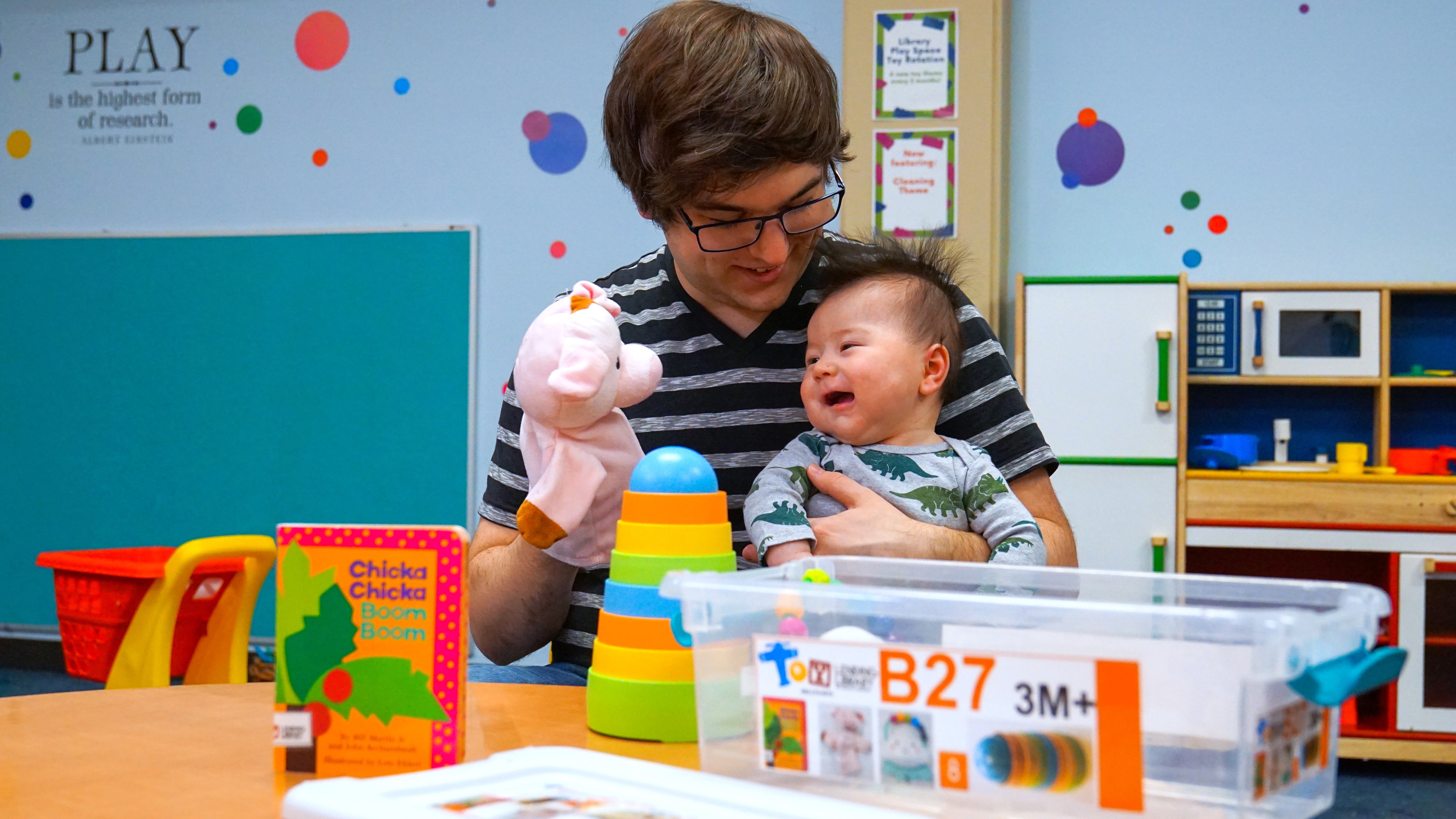 In the Downtown Library's play room, a father makes his baby laugh with a hand puppet. An open toy box and a board book are on the table in front of them.