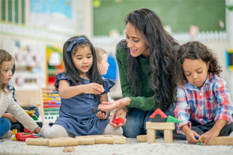 A woman helps three small children as they play with wooden blocks.