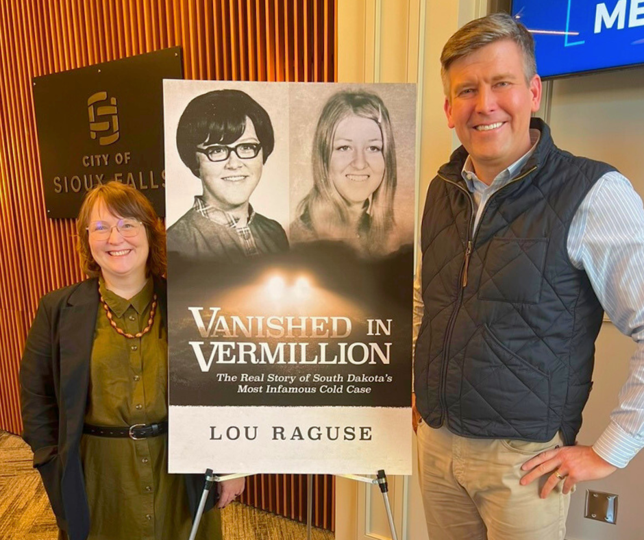 Lou Raguse and Kim Koblank standing next to a poster size book cover of Vanished in Vermillion for the One Book Siouxland title announcement.